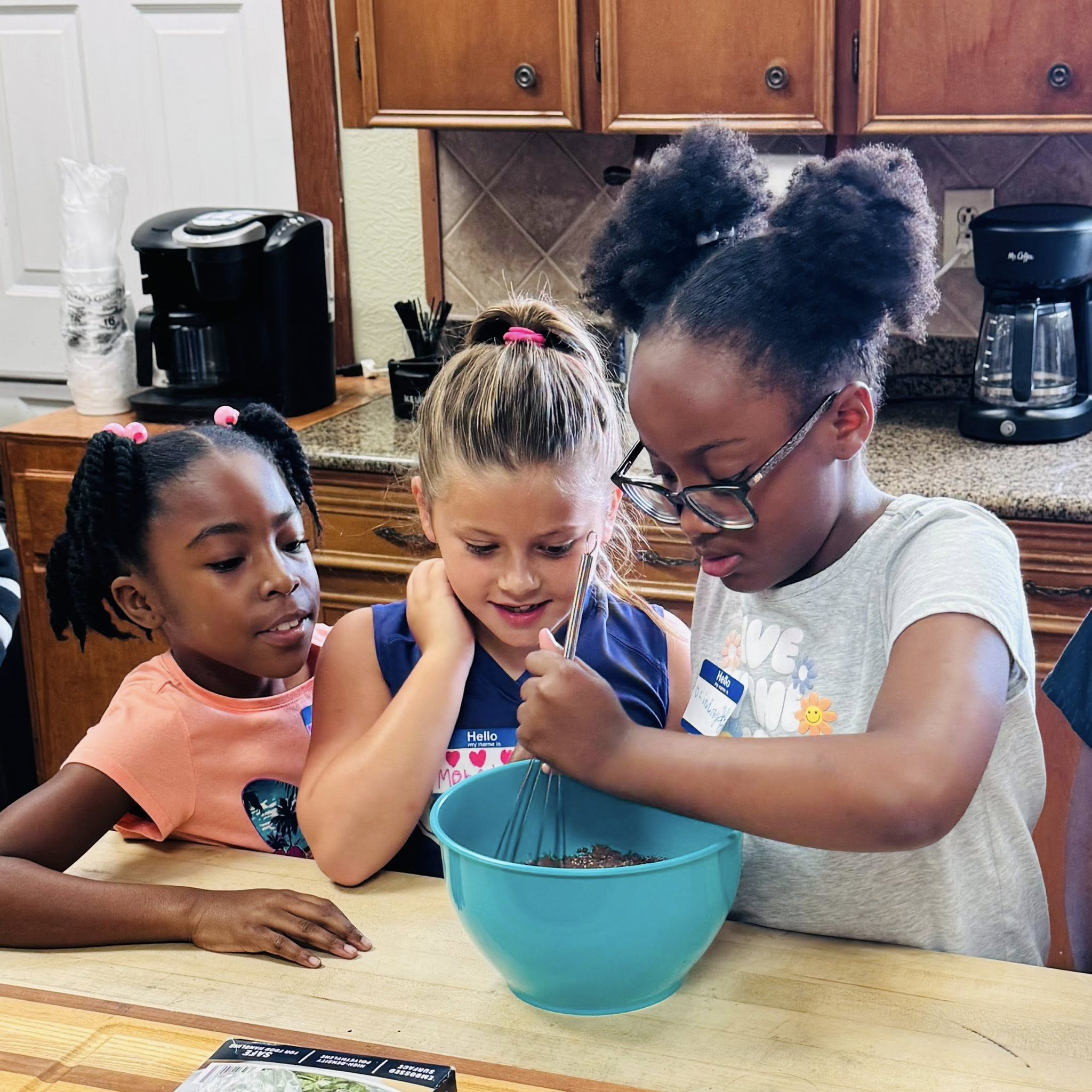Two girls watching another girl whisk in a bowl.